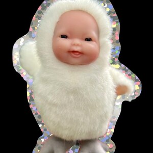 May include: A small doll in a white, furry hooded outfit with matching mittens and grey boots. The doll has a smiling face and is set against a black background with a decorative border.