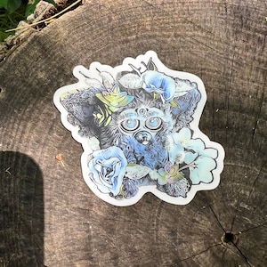 May include: A blue and white sticker featuring a cartoon creature with large eyes and a floral design. The creature is surrounded by flowers and leaves.