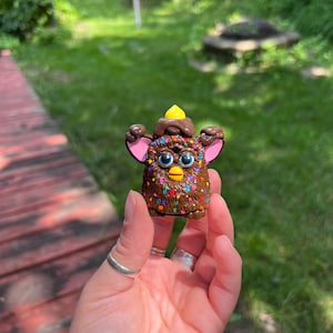 May include: A small, brown Furby-like toy with colourful sprinkles, blue eyes, and pink ears. It has a yellow top and is held in a hand with silver rings. The background shows green grass and trees.