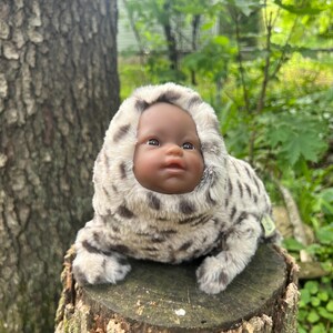 May include: A doll with dark skin wearing a plush seal costume with a hood. The costume is light grey with dark grey spots. The doll is sitting on a tree stump in a natural outdoor setting.