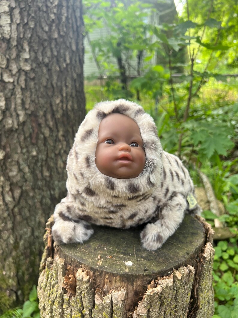 May include: A doll with dark skin wearing a plush seal costume with a hood. The costume is light grey with dark grey spots. The doll is sitting on a tree stump in a natural outdoor setting.