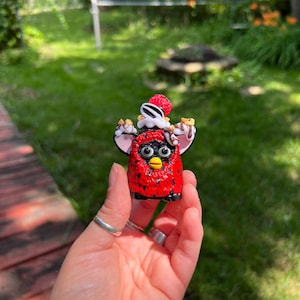 May include: A red Furby figurine with black spots, yellow eyes, and a dessert-themed hat. The figurine is held in a hand with silver rings. The background is a blurred outdoor scene with green grass and trees.