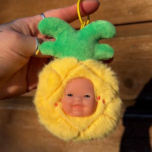 May include: A plush pineapple-shaped keychain with a baby doll face. The pineapple has a yellow, fuzzy body with a green, plush crown. The baby doll face is centered in the pineapple, with a smiling expression. A yellow loop is attached for hanging.