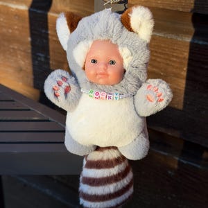 May include: A plush raccoon-themed keychain featuring a doll face. The keychain has a gray and white body, brown ears, and a striped tail. The doll has blue eyes and a necklace that spells "ROCKY".