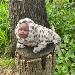 May include: A plush doll with a dark complexion and a hooded, spotted fur suit, resembling a seal. The doll is resting on a tree stump in a natural outdoor setting, surrounded by greenery.