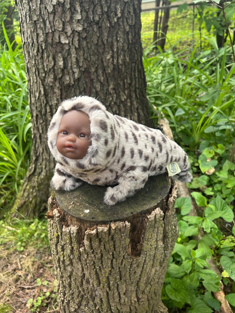 May include: A plush doll with a dark complexion and a hooded, spotted fur suit, resembling a seal. The doll is resting on a tree stump in a natural outdoor setting, surrounded by greenery.