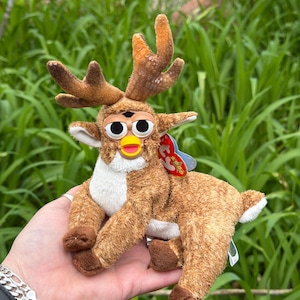 Könnte beinhalten: Ein braunes Plüschtier mit Hirschgeweih und einem Furby-ähnlichen Gesicht. Das Spielzeug hat eine weiße Brust, braune Beine und ein rot-blaues Etikett. Der Hintergrund ist verschwommenes grünes Laub.