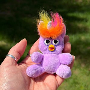 May include: A small, plush purple creature with large, round eyes and a yellow beak. It has a colorful, rainbow-coloured tuft of hair and a small, white horn. The toy is held in a hand against a blurred green background.