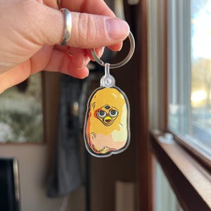May include: A yellow and orange keychain featuring a cartoon character that looks like a Furby. The character has big eyes, a small beak, and a fluffy texture. The keychain is attached to a silver keyring.