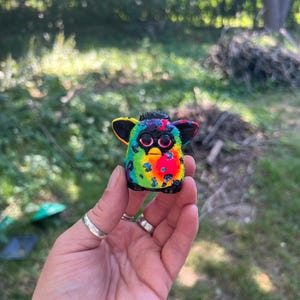 May include: A small Furby figurine with a vibrant rainbow leopard print pattern. It has black ears and a black and purple top. The eyes are pink. The figurine is held in a hand.
