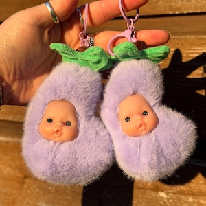 Könnte beinhalten: Zwei Schlüsselanhänger mit einer flauschigen, lavendelfarbenen Birnenform und einem Babypuppengesicht. Jeder Schlüsselanhänger hat ein grünes Filzblatt und einen rosafarbenen Metallverschluss. Die Schlüsselanhänger werden vor einem Holzgrund gezeigt.