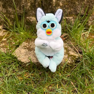 May include: A Furby plush toy with a light blue body, white details, and black and white ears. The toy has large eyes, a yellow beak, and a red mouth. It is positioned on a stone surface, surrounded by grass.