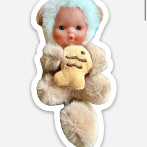 May include: A doll in a fluffy brown outfit with a white fur-trimmed hood. The doll is holding a small, yellow plush toy with a wavy design. The doll has a realistic face with blue eyes and rosy cheeks.