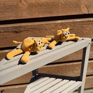 May include: Two plush, yellow toy creatures with large, expressive eyes and outstretched limbs are displayed on a gray shelf. The toys have a whimsical design, with a soft, textured appearance. The background features a wooden wall.