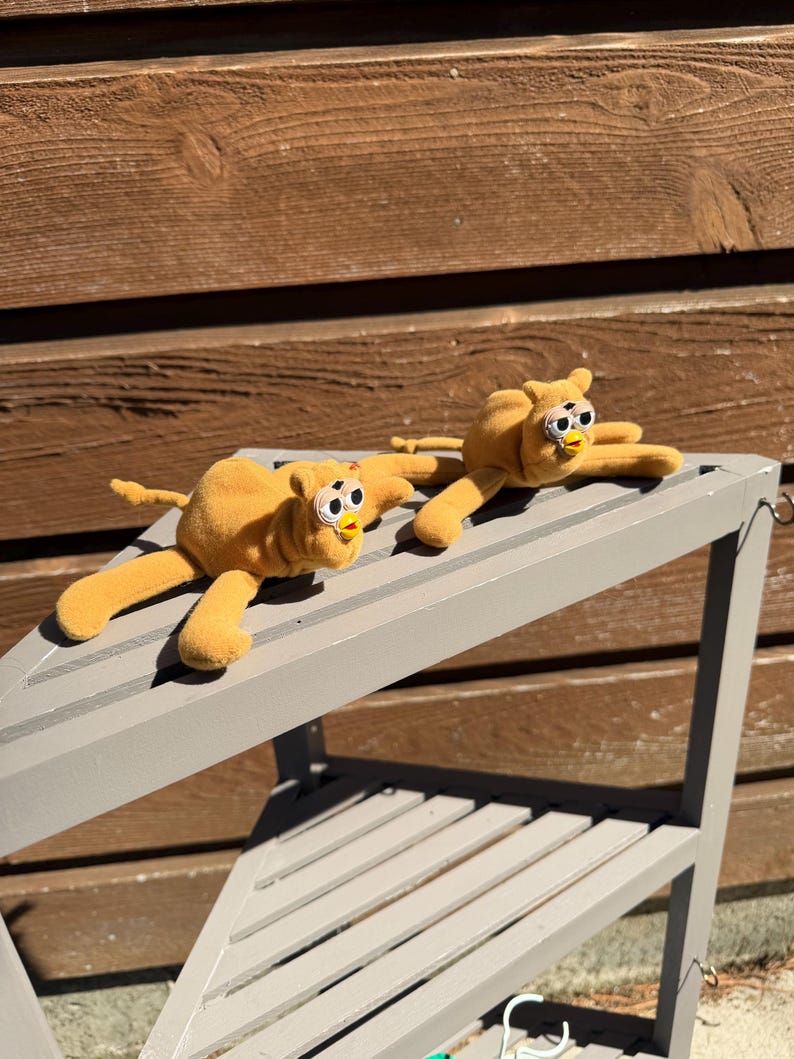 May include: Two plush, yellow toy creatures with large, expressive eyes and outstretched limbs are displayed on a gray shelf. The toys have a whimsical design, with a soft, textured appearance. The background features a wooden wall.