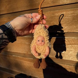 May include: A handmade keychain featuring a small doll dressed in a fuzzy, tan outfit with brown fabric legs. The doll has a light skin tone and is attached to a brown braided cord.
