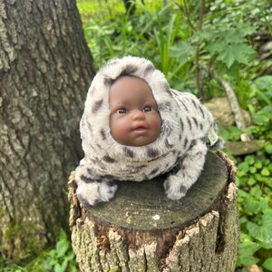 May include: A baby doll with dark skin and brown eyes, wearing a plush, hooded outfit with a spotted pattern in shades of grey and cream. The doll is posed on a weathered tree stump, surrounded by greenery.
