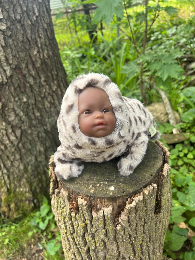 May include: A baby doll with dark skin and brown eyes, wearing a plush, hooded outfit with a spotted pattern in shades of grey and cream. The doll is posed on a weathered tree stump, surrounded by greenery.