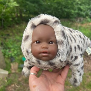 May include: A seal-shaped plush toy with a baby doll face. The seal's fur is white and grey, with a spotted pattern. The doll has brown eyes and a neutral expression. The toy is held in a hand against a blurred green background.