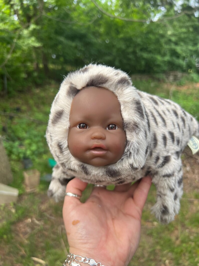 May include: A seal-shaped plush toy with a baby doll face. The seal's fur is white and grey, with a spotted pattern. The doll has brown eyes and a neutral expression. The toy is held in a hand against a blurred green background.