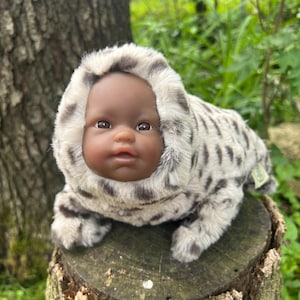 May include: A baby doll with dark skin and brown eyes in a plush, hooded outfit with a spotted pattern in grey and white. The doll is posed on a tree stump outdoors.