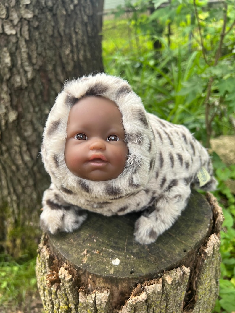 May include: A baby doll with dark skin and brown eyes in a plush, hooded outfit with a spotted pattern in grey and white. The doll is posed on a tree stump outdoors.