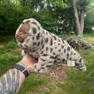 May include: A seal-shaped plush toy with a dark-skinned baby doll face. The toy's body is patterned with grey and brown spots. The toy is held up against a backdrop of green foliage. The toy has a small tag attached.