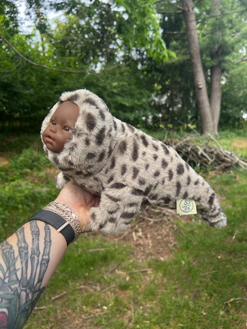 May include: A seal-shaped plush toy with a dark-skinned baby doll face. The toy's body is patterned with grey and brown spots. The toy is held up against a backdrop of green foliage. The toy has a small tag attached.