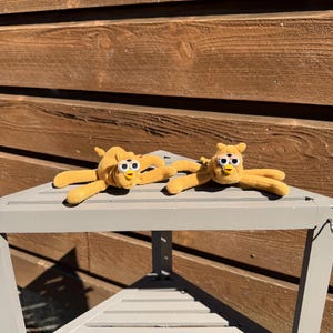 May include: Two plush, yellow creatures with large, white-rimmed eyes and small beaks are lying on a gray, wooden table. The creatures have long, slender arms and legs. The background is a wooden wall.