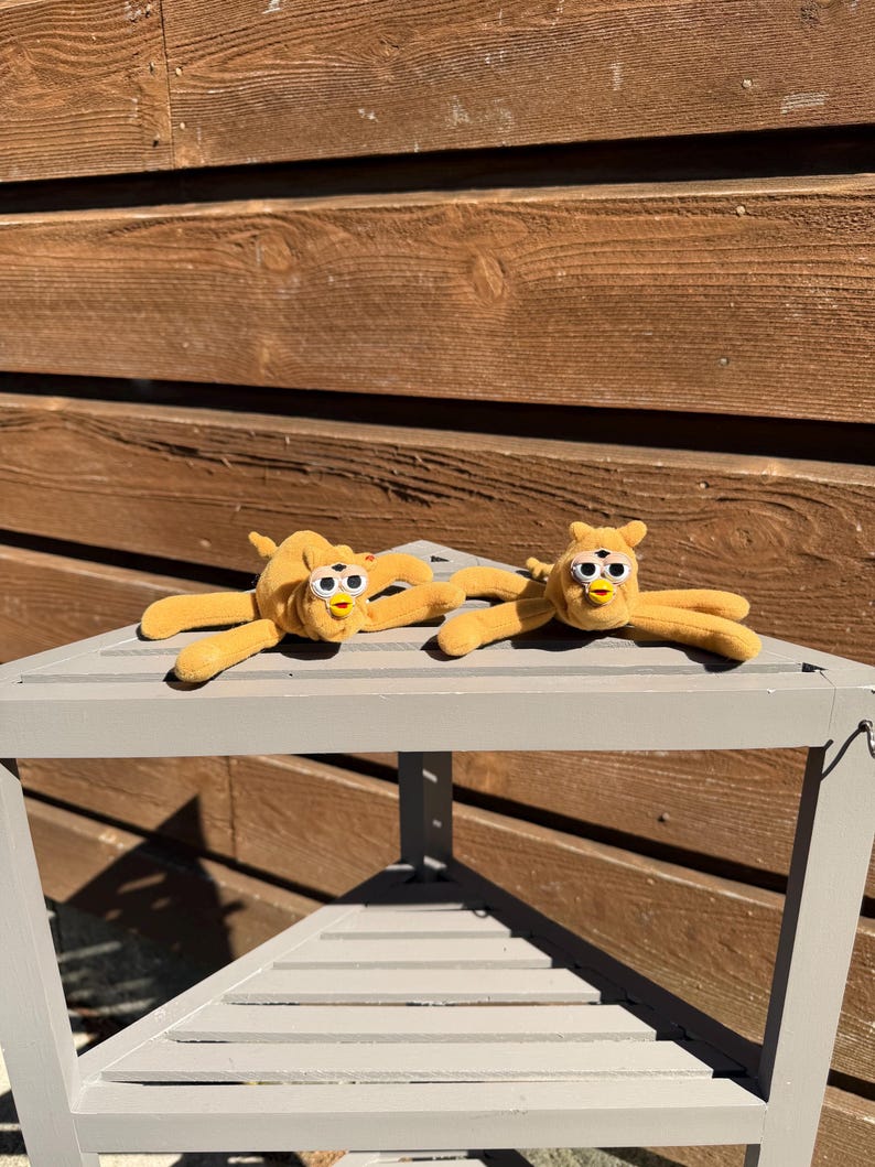 May include: Two plush, yellow creatures with large, white-rimmed eyes and small beaks are lying on a gray, wooden table. The creatures have long, slender arms and legs. The background is a wooden wall.
