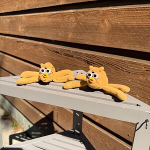 May include: Two plush, yellow Furby toys with open arms and round eyes are displayed on a gray bench. The toys have a classic design with a beak and small, yellow arms and legs. The background features a wooden wall.
