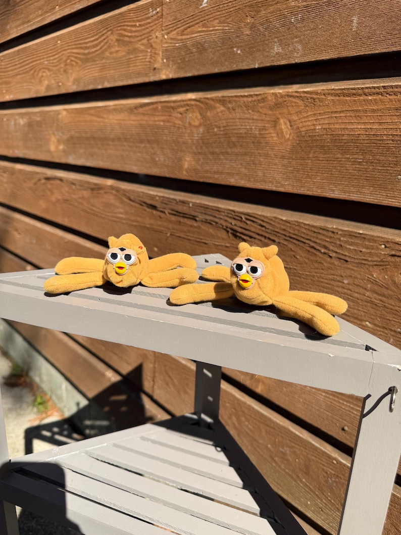 May include: Two plush, yellow Furby toys with open arms and round eyes are displayed on a gray bench. The toys have a classic design with a beak and small, yellow arms and legs. The background features a wooden wall.