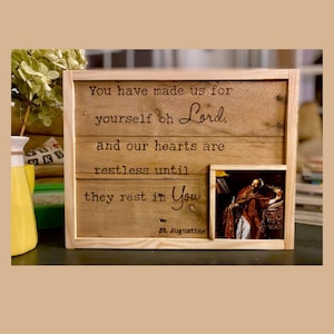 May include: A rectangular wooden sign with a light-colored frame featuring the engraved text: You have made us for yourself oh Lord, and our hearts are restless until they rest in You. St. Augustine. A small color image of a figure is inset.