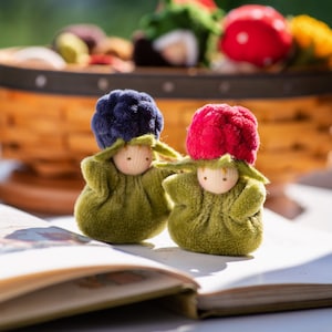 Pode incluir: Duas pequenas bonecas de feltro verde com rostos bordados. As bonecas têm chapéus de feltro de cores diferentes, uma é azul e a outra é vermelha. As bonecas estão sentadas em um livro.