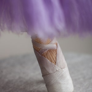 May include: A close-up of a white fabric ballerina shoe with a purple tulle skirt in the background. The shoe has a white ribbon tied around it.