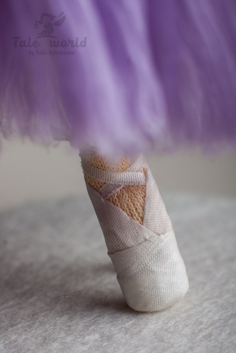 May include: A close-up of a white fabric ballerina shoe with a purple tulle skirt in the background. The shoe has a white ribbon tied around it.