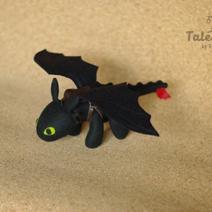 May include: Black felt plush toy dragon with green eyes and a leather saddle. The dragon has wings and a tail. The toy is on a brown background.