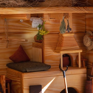 Puede incluir: Un interior de casa de muñecas de madera en miniatura con una chimenea, una cama pequeña y un suelo de madera. La chimenea tiene una olla pequeña y un atizador de metal. La cama tiene un cojín rojo y verde. Hay hierbas secas colgando del techo.