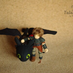 May include: A black and green plush dragon with wings, a small doll with brown hair wearing a dark green and brown outfit is riding on the dragon's back. The dragon is looking to the left. The doll is looking down at the dragon. The background is a light brown surface.