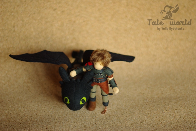 May include: A black and green plush dragon with wings, a small doll with brown hair wearing a dark green and brown outfit is riding on the dragon's back. The dragon is looking to the left. The doll is looking down at the dragon. The background is a light brown surface.