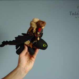 May include: A black dragon plush toy with a green eye, holding a small doll with brown hair and a brown fur coat. The dragon is flying with its wings spread out.