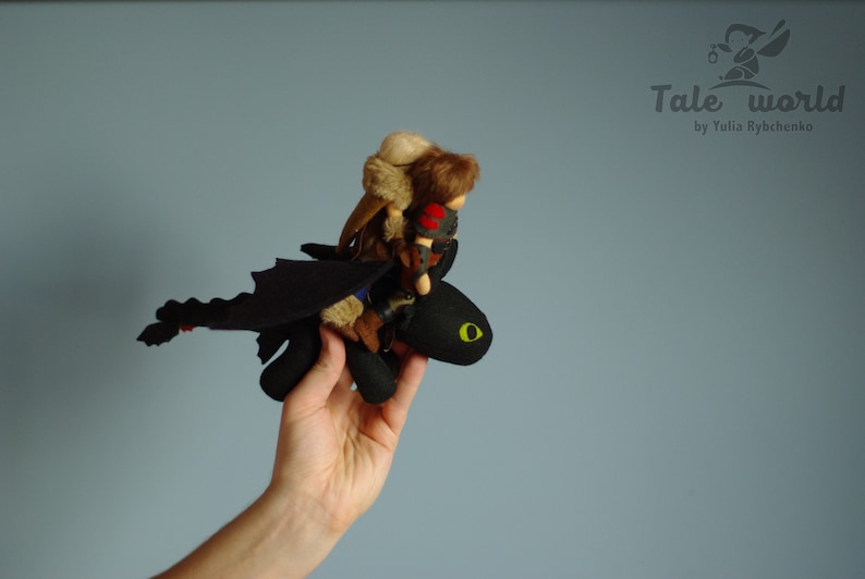 May include: A black dragon plush toy with a green eye, holding a small doll with brown hair and a brown fur coat. The dragon is flying with its wings spread out.