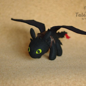 May include: Black plush toy dragon with green eyes and a brown leather saddle. The dragon has wings and a tail. The toy is on a brown background.