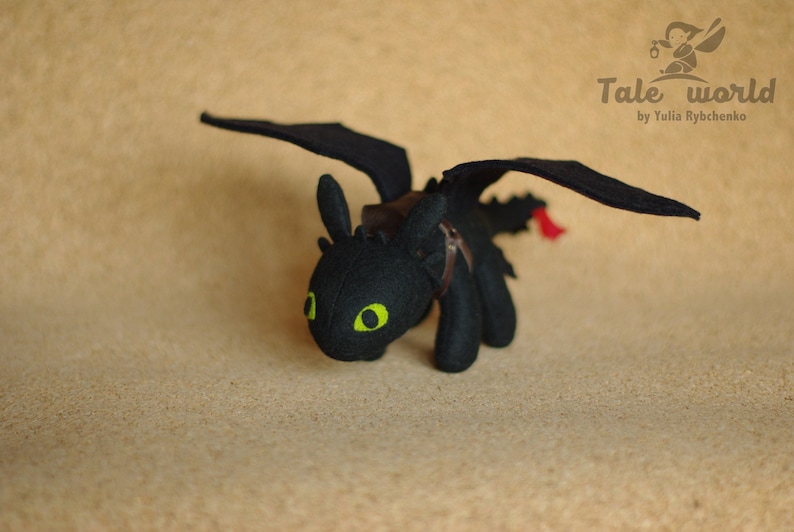 May include: Black plush toy dragon with green eyes and a brown leather saddle. The dragon has wings and a tail. The toy is on a brown background.