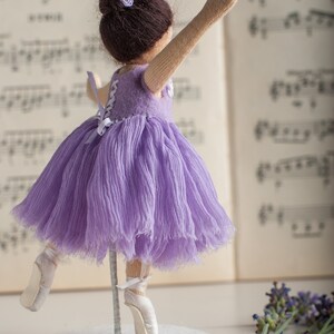 May include: A purple ballerina doll with a white tutu and white ballet shoes. The doll is standing on a white base with a beaded edge. The doll is in front of a sheet of music and a bouquet of lavender.
