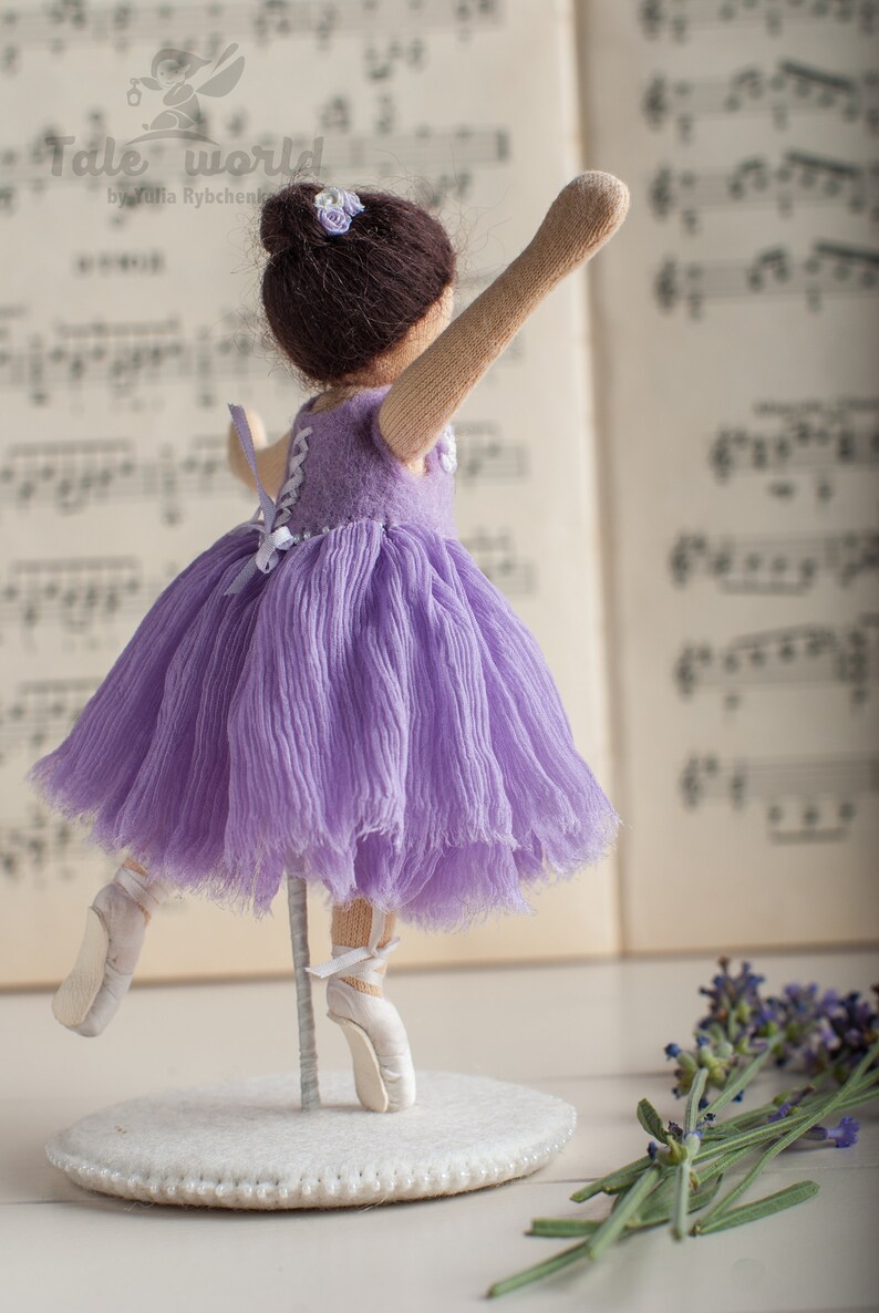 May include: A purple ballerina doll with a white tutu and white ballet shoes. The doll is standing on a white base with a beaded edge. The doll is in front of a sheet of music and a bouquet of lavender.