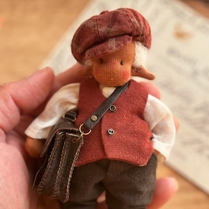 May include: A handmade doll dressed in a red vest, cap, and dark pants, carrying a small brown satchel. The doll has a light skin tone and white hair. The doll is a miniature figure.