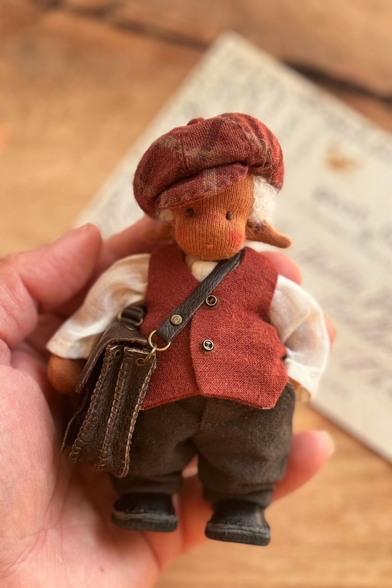 May include: A handmade doll dressed in a red vest, cap, and dark pants, carrying a small brown satchel. The doll has a light skin tone and white hair. The doll is a miniature figure.