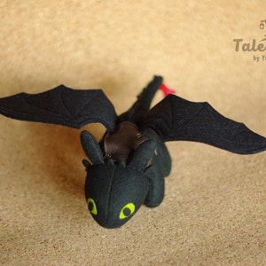 May include: Black felt plush toy dragon with green eyes and red accents. The dragon has wings and a tail. The toy is on a brown background.