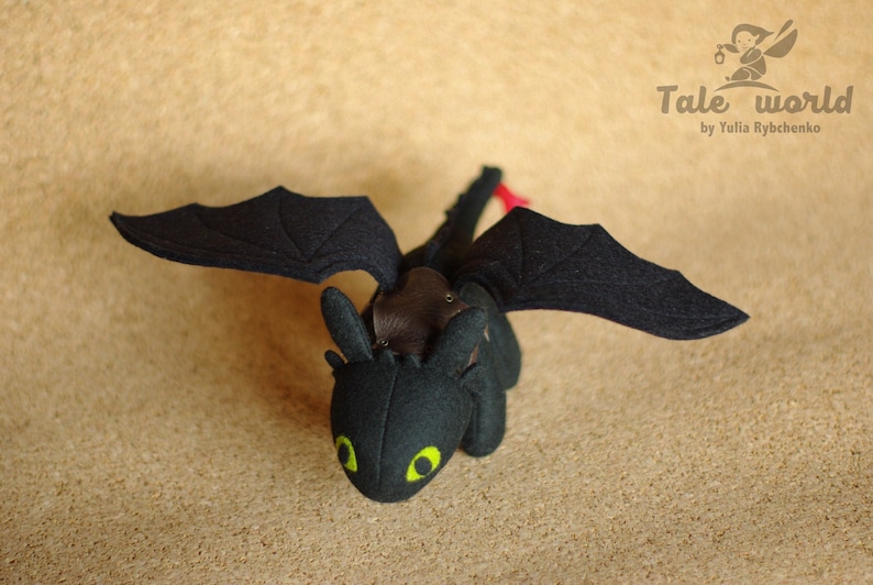May include: Black felt plush toy dragon with green eyes and red accents. The dragon has wings and a tail. The toy is on a brown background.